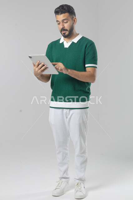استعمال التقنية الحديثه المتطورة في الحياة العملية عبر الجهاز اللوحي، تعابير وايماءات الاندماج، بورتريه لشاب عربي خليجي سعودي مبتسم يرتدي الزي الكاجوال يقوم بالعمل على جهازالتابلت، صورة الجسم بالطول الكامل، خلفية بيضاء