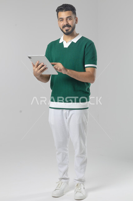 استعمال التقنية الحديثه المتطورة في الحياة العملية عبر الجهاز اللوحي، تعابير وايماءات الاندماج، بورتريه لشاب عربي خليجي سعودي مبتسم يرتدي الزي الكاجوال يقوم بالعمل على جهازالتابلت، صورة الجسم بالطول الكامل، خلفية بيضاء