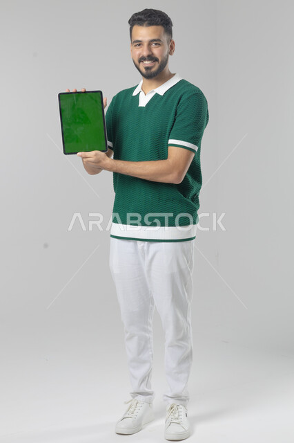 عرض شاشة خضراء كروما عبر الجهاز اللوحي، استعمال التقنية الحديثه المتطورة في الحياة العملية، تعابير وايماءات الاندماج، بورتريه لشاب عربي خليجي سعودي مبتسم يرتدي الزي الكاجوال يقوم بالعمل على جهازالتابلت، صورة الجسم بالطول الكامل، خلفية بيضاء