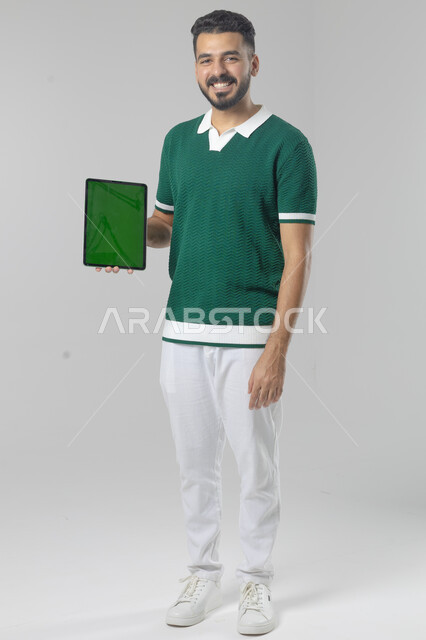 عرض شاشة خضراء كروما عبر الجهاز اللوحي، استعمال التقنية الحديثه المتطورة في الحياة العملية، تعابير وايماءات الاندماج، بورتريه لشاب عربي خليجي سعودي مبتسم يرتدي الزي الكاجوال يقوم بالعمل على جهازالتابلت، صورة الجسم بالطول الكامل، خلفية بيضاء