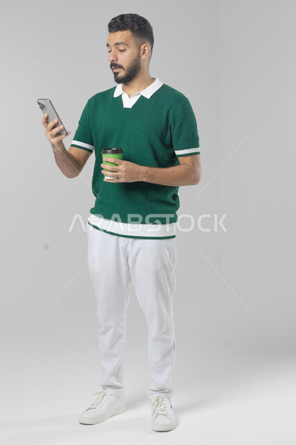 Eating and drinking hot drinks, using modern advanced technology in practical life, portrait of a smiling Saudi Arabian Gulf young man wearing casual clothes working on a mobile phone, full body image, white background