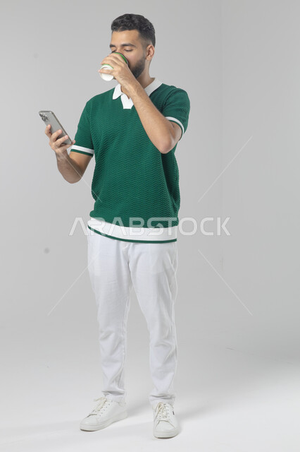 Eating and drinking hot drinks, using modern advanced technology in practical life, portrait of a smiling Saudi Arabian Gulf young man wearing casual clothes working on a mobile phone, full body image, white background