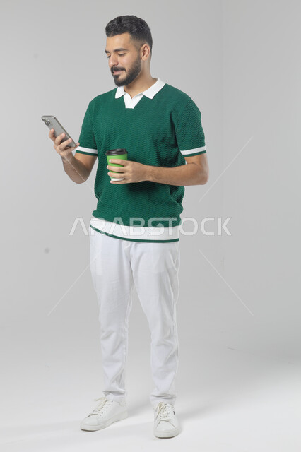 Eating and drinking hot drinks, using modern advanced technology in practical life, portrait of a smiling Saudi Arabian Gulf young man wearing casual clothes working on a mobile phone, full body image, white background