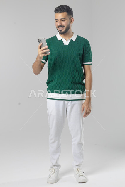  تعابير وايماءات الاندماج والسرور، استخدام التقنية الحديثه المتطورة في الحياة العملية، بورتريه لشاب عربي خليجي سعودي مبتسم يرتدي الزي الكاجوال يقوم بالعمل على الهاتف المحمول، صورة الجسم بالطول الكامل، خلفية بيضاء