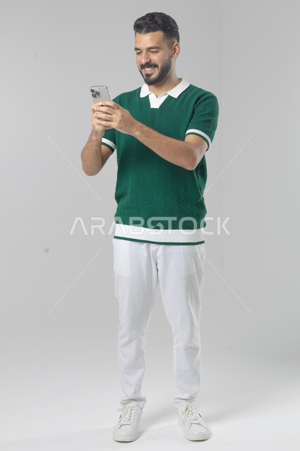  تعابير وايماءات الاندماج والسرور، استخدام التقنية الحديثه المتطورة في الحياة العملية، بورتريه لشاب عربي خليجي سعودي مبتسم يرتدي الزي الكاجوال يقوم بالعمل على الهاتف المحمول، صورة الجسم بالطول الكامل، خلفية بيضاء
