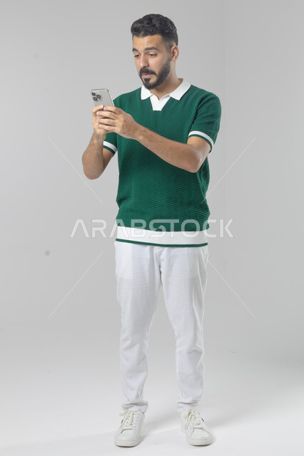  تعابير وايماءات الاندماج والسرور، استخدام التقنية الحديثه المتطورة في الحياة العملية، بورتريه لشاب عربي خليجي سعودي مبتسم يرتدي الزي الكاجوال يقوم بالعمل على الهاتف المحمول، صورة الجسم بالطول الكامل، خلفية بيضاء