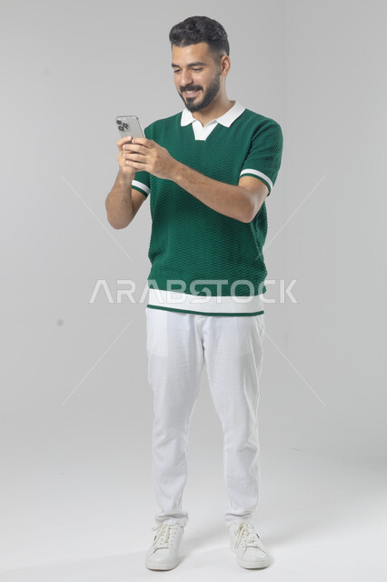  تعابير وايماءات الاندماج والسرور، استخدام التقنية الحديثه المتطورة في الحياة العملية، بورتريه لشاب عربي خليجي سعودي مبتسم يرتدي الزي الكاجوال يقوم بالعمل على الهاتف المحمول، صورة الجسم بالطول الكامل، خلفية بيضاء