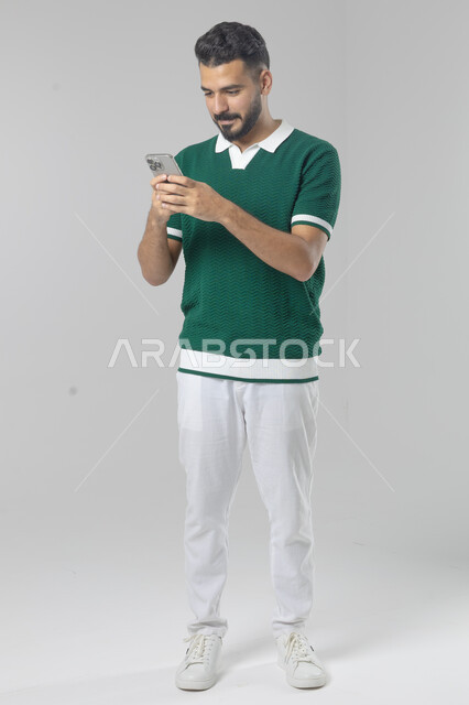  تعابير وايماءات الاندماج والسرور، استخدام التقنية الحديثه المتطورة في الحياة العملية، بورتريه لشاب عربي خليجي سعودي مبتسم يرتدي الزي الكاجوال يقوم بالعمل على الهاتف المحمول، صورة الجسم بالطول الكامل، خلفية بيضاء