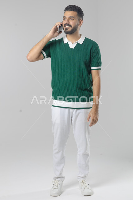 Chatting and conversing using a modern technical device, answering a phone call, portrait of a Saudi Arabian Gulf man in casual wear making a phone call through a mobile phone, full body image, white background