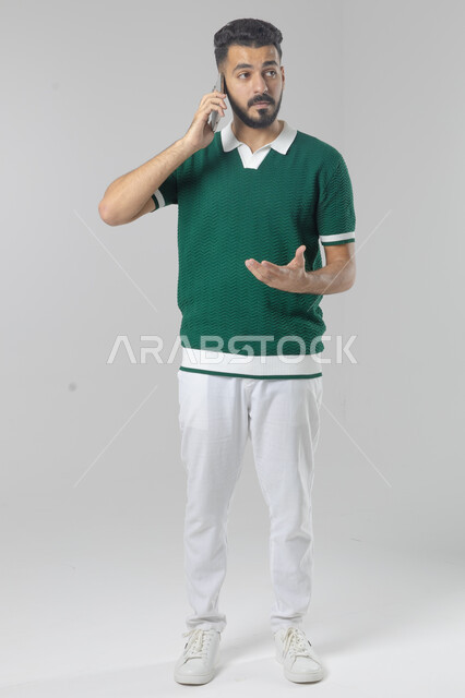 Chatting and conversing using a modern technical device, answering a phone call, portrait of a Saudi Arabian Gulf man in casual wear making a phone call through a mobile phone, full body image, white background