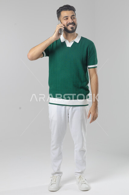Chatting and conversing using a modern technical device, answering a phone call, portrait of a Saudi Arabian Gulf man in casual wear making a phone call through a mobile phone, full body image, white background