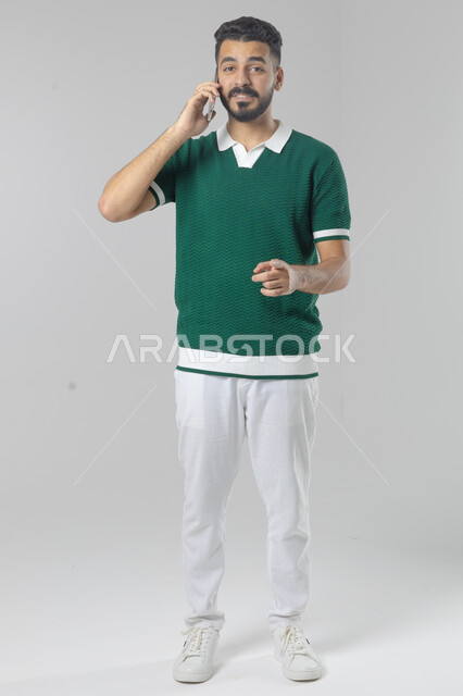 Chatting and conversing using a modern technical device, answering a phone call, portrait of a Saudi Arabian Gulf man in casual wear making a phone call through a mobile phone, full body image, white background