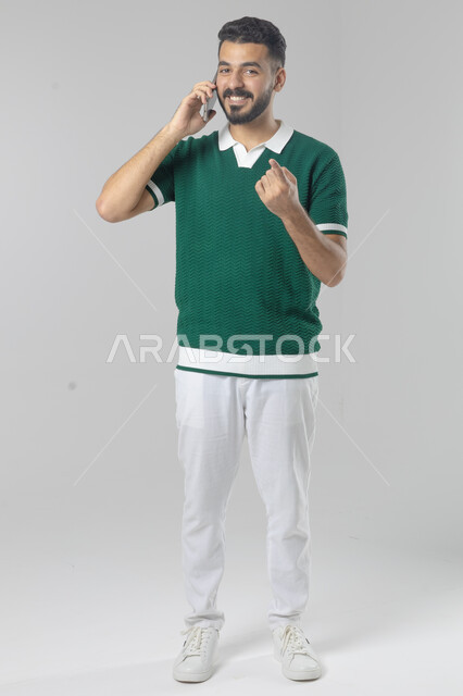 الدردشة والمحادثة باستخدام جهاز تقني حديث, الرد على مكالمة هاتفية، بورتريه لرجل عربي خليجي سعودي يرتدي الزي الكاجوال يقوم باجراء اتصال هاتفي من خلال الهاتف المحمول، صورة الجسم بالطول الكامل, خلفية بيضاء