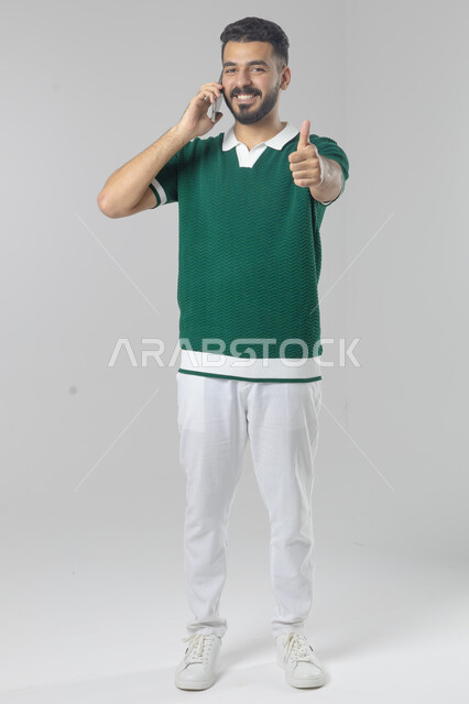 Chatting and conversing using a modern technical device, answering a phone call, portrait of a Saudi Arabian Gulf man in casual wear making a phone call through a mobile phone, full body image, white background