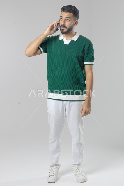 Chatting and conversing using a modern technical device, answering a phone call, portrait of a Saudi Arabian Gulf man in casual wear making a phone call through a mobile phone, full body image, white background