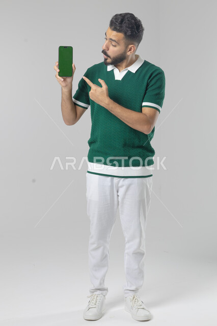  تعابير وايماءات الاندماج والسرور، استخدام التقنية الحديثه المتطورة في الحياة العملية، بورتريه لشاب عربي خليجي سعودي مبتسم يرتدي الزي الكاجوال يقوم بعرض شاشة فارغة على الهاتف المحمول، صورة الجسم بالطول الكامل، خلفية بيضاء