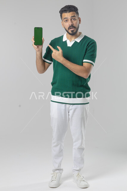  تعابير وايماءات الاندماج والسرور، استخدام التقنية الحديثه المتطورة في الحياة العملية، بورتريه لشاب عربي خليجي سعودي مبتسم يرتدي الزي الكاجوال يقوم بعرض شاشة فارغة على الهاتف المحمول، صورة الجسم بالطول الكامل، خلفية بيضاء