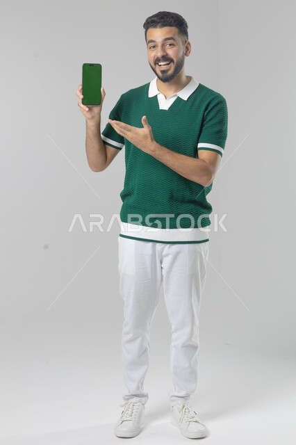  تعابير وايماءات الاندماج والسرور، استخدام التقنية الحديثه المتطورة في الحياة العملية، بورتريه لشاب عربي خليجي سعودي مبتسم يرتدي الزي الكاجوال يقوم بعرض شاشة فارغة على الهاتف المحمول، صورة الجسم بالطول الكامل، خلفية بيضاء
