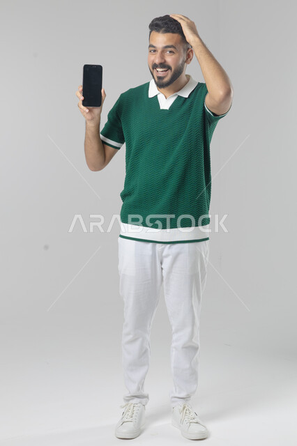  تعابير وايماءات الاندماج والسرور، استخدام التقنية الحديثه المتطورة في الحياة العملية، بورتريه لشاب عربي خليجي سعودي مبتسم يرتدي الزي الكاجوال يقوم بعرض شاشة فارغة على الهاتف المحمول، صورة الجسم بالطول الكامل، خلفية بيضاء