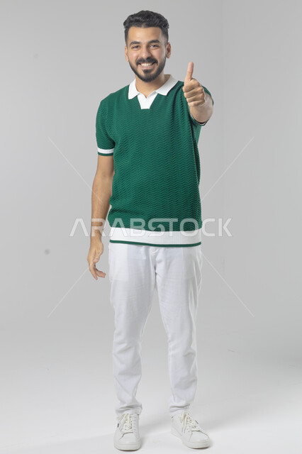حركة أصبع تدل على القبول والرضا، الإمتياز والتميز والنجاح، بورتريه لرجل عربي خليجي سعودي يرتدي الزي الكاجوال ويرفع أصابع الإبهام لأعلى، إيماءات السعادة والإعجاب والموافقة، الجسم بالطول الكامل، خلفية بيضاء 