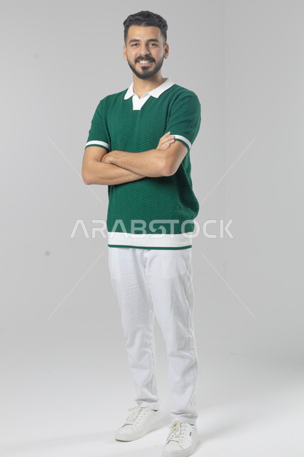  التقاط صور شخصية للوثائق الرسمية، الاعتناء والاهتمام بالمظهر الخارجي، مفهوم الاناقة والرجولة، بورتريه لشاب عربي خليجي سعودي يرتدي الزي الكاجوال يقف بأيدي متقاطعة ينظر الى الكاميرا بتعابير السرور والثقة بالنفس، الجسم بالطول الكامل، خلفية بيضاء