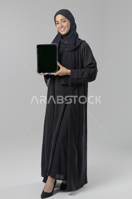  استخدام التقنية الحديثه المتطورة في الحياة العملية، عرض الشاشة عبر الجهاز اللوحي، تعابير وايماءات الاندماج، بورتريه لشابة عربية خليجية سعودية مبتسمة ترتدي الحجاب والعباءة تقوم بالعمل على جهازالتابلت، صورة الجسم بالطول الكامل، خلفية بيضاء 