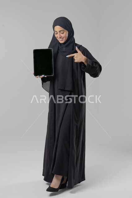 Using modern advanced technology in practical life, displaying the screen via a tablet, expressions and gestures of integration, portrait of a smiling young Saudi Arabian Gulf woman wearing a hijab and abaya working on a tablet, full-length body image, white background