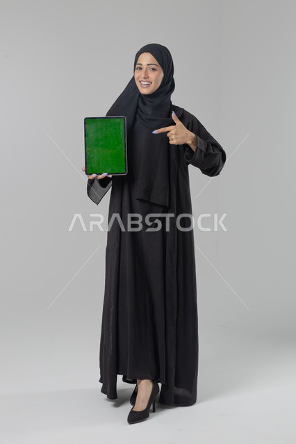  استخدام التقنية الحديثه المتطورة في الحياة العملية، عرض الشاشة عبر الجهاز اللوحي، تعابير وايماءات الاندماج، بورتريه لشابة عربية خليجية سعودية مبتسمة ترتدي الحجاب والعباءة تقوم بالعمل على جهازالتابلت، صورة الجسم بالطول الكامل، خلفية بيضاء 