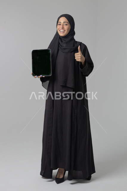  استخدام التقنية الحديثه المتطورة في الحياة العملية، عرض الشاشة عبر الجهاز اللوحي، تعابير وايماءات الاندماج، بورتريه لشابة عربية خليجية سعودية مبتسمة ترتدي الحجاب والعباءة تقوم بالعمل على جهازالتابلت، صورة الجسم بالطول الكامل، خلفية بيضاء 