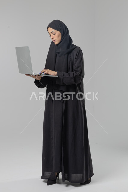  مهن ووظائف نسائية سعودية، ادارة الاعمال عبر التقنية والأجهزة الحديثة، بورتريه لإمرأة عربية خليجية سعودية مبتسمة ترتدي العباءة والحجاب تمسك الحاسب المحمول، التسويق الالكتروني الذكي والآمن، صورة الجسم بالكامل، خلفية بيضاء