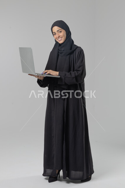  مهن ووظائف نسائية سعودية، ادارة الاعمال عبر التقنية والأجهزة الحديثة، بورتريه لإمرأة عربية خليجية سعودية مبتسمة ترتدي العباءة والحجاب تمسك الحاسب المحمول، التسويق الالكتروني الذكي والآمن، صورة الجسم بالكامل، خلفية بيضاء