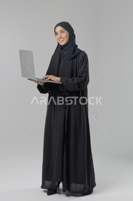  مهن ووظائف نسائية سعودية، ادارة الاعمال عبر التقنية والأجهزة الحديثة، بورتريه لإمرأة عربية خليجية سعودية مبتسمة ترتدي العباءة والحجاب تمسك الحاسب المحمول، التسويق الالكتروني الذكي والآمن، صورة الجسم بالكامل، خلفية بيضاء