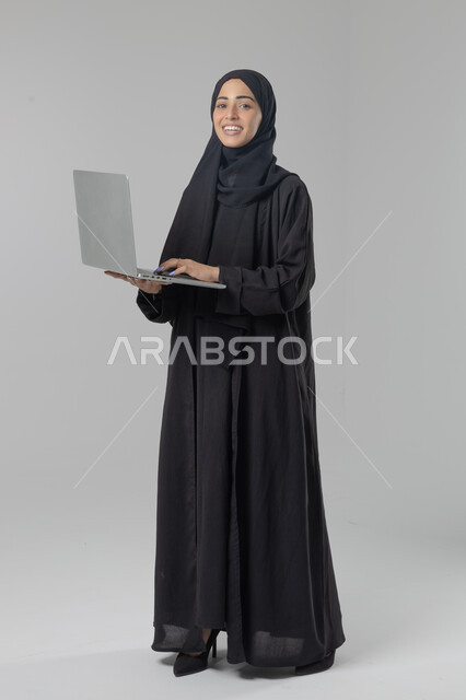  مهن ووظائف نسائية سعودية، ادارة الاعمال عبر التقنية والأجهزة الحديثة، بورتريه لإمرأة عربية خليجية سعودية مبتسمة ترتدي العباءة والحجاب تمسك الحاسب المحمول، التسويق الالكتروني الذكي والآمن، صورة الجسم بالكامل، خلفية بيضاء