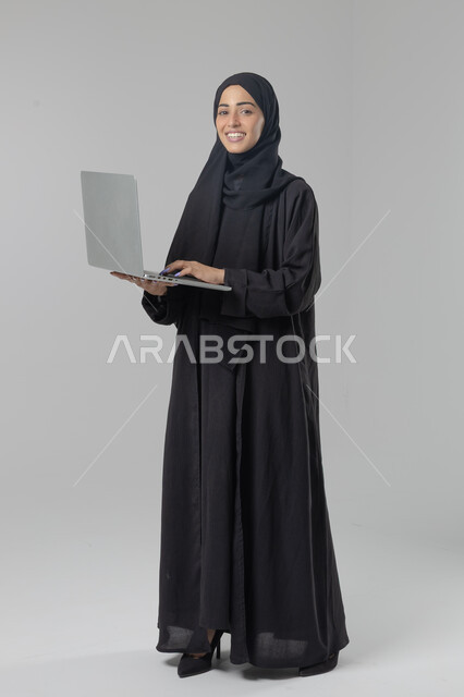  مهن ووظائف نسائية سعودية، ادارة الاعمال عبر التقنية والأجهزة الحديثة، بورتريه لإمرأة عربية خليجية سعودية مبتسمة ترتدي العباءة والحجاب تمسك الحاسب المحمول، التسويق الالكتروني الذكي والآمن، صورة الجسم بالكامل، خلفية بيضاء