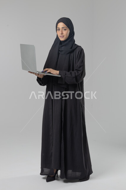  مهن ووظائف نسائية سعودية، ادارة الاعمال عبر التقنية والأجهزة الحديثة، بورتريه لإمرأة عربية خليجية سعودية مبتسمة ترتدي العباءة والحجاب تمسك الحاسب المحمول، التسويق الالكتروني الذكي والآمن، صورة الجسم بالكامل، خلفية بيضاء