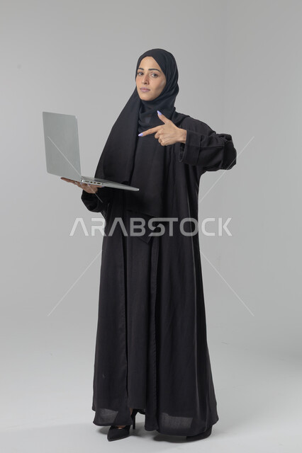  مهن ووظائف نسائية سعودية، ادارة الاعمال عبر التقنية والأجهزة الحديثة، بورتريه لإمرأة عربية خليجية سعودية مبتسمة ترتدي العباءة والحجاب تمسك الحاسب المحمول، التسويق الالكتروني الذكي والآمن، صورة الجسم بالكامل، خلفية بيضاء