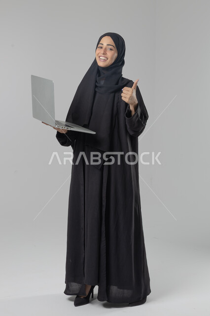  مهن ووظائف نسائية سعودية، ادارة الاعمال عبر التقنية والأجهزة الحديثة، بورتريه لإمرأة عربية خليجية سعودية مبتسمة ترتدي العباءة والحجاب تمسك الحاسب المحمول، التسويق الالكتروني الذكي والآمن، صورة الجسم بالكامل، خلفية بيضاء