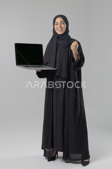  مهن ووظائف نسائية سعودية، ادارة الاعمال عبر التقنية والأجهزة الحديثة، بورتريه لإمرأة عربية خليجية سعودية مبتسمة ترتدي العباءة والحجاب تمسك الحاسب المحمول، التسويق الالكتروني الذكي والآمن، صورة الجسم بالكامل، خلفية بيضاء