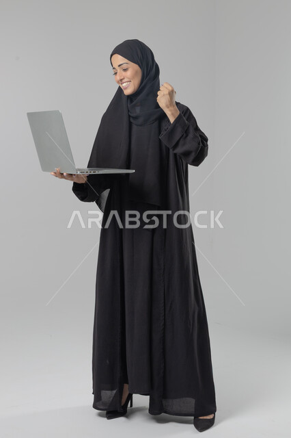  مهن ووظائف نسائية سعودية، ادارة الاعمال عبر التقنية والأجهزة الحديثة، بورتريه لإمرأة عربية خليجية سعودية مبتسمة ترتدي العباءة والحجاب تمسك الحاسب المحمول، التسويق الالكتروني الذكي والآمن، صورة الجسم بالكامل، خلفية بيضاء