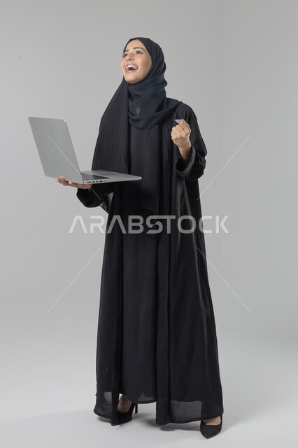  مهن ووظائف نسائية سعودية، ادارة الاعمال عبر التقنية والأجهزة الحديثة، بورتريه لإمرأة عربية خليجية سعودية مبتسمة ترتدي العباءة والحجاب تمسك الحاسب المحمول، التسويق الالكتروني الذكي والآمن، صورة الجسم بالكامل، خلفية بيضاء