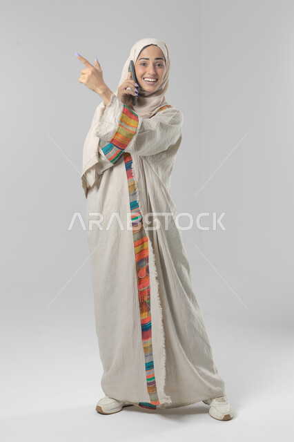 التحدث عبر الهاتف المحمول، قضاء اوقات ممتعة بتبادل الاخبار والأحاديث، بورتريه لامرأة عربية خليجية سعودية محجبة ترتدي العباءة تقوم بإجرء مكالمة هاتفية، محادثات ودردشات مع الاصدقاء، صورة الجسم بالكامل، خلفية بيضاء