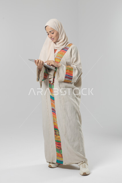 Using modern advanced technology in practical life via tablet, expressions and gestures of integration, portrait of a smiling young Saudi Arabian Gulf woman wearing hijab and abaya working on a tablet, full body image, white background