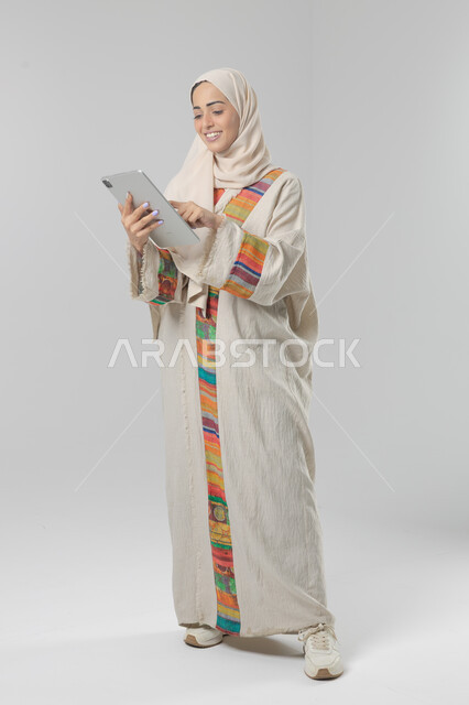  استخدام التقنية الحديثه المتطورة في الحياة العملية عبر الجهاز اللوحي، تعابير وايماءات الاندماج، بورتريه لشابة عربية خليجية سعودية مبتسمة ترتدي الحجاب والعباءة تقوم بالعمل على جهازالتابلت، صورة الجسم بالطول الكامل، خلفية بيضاء 