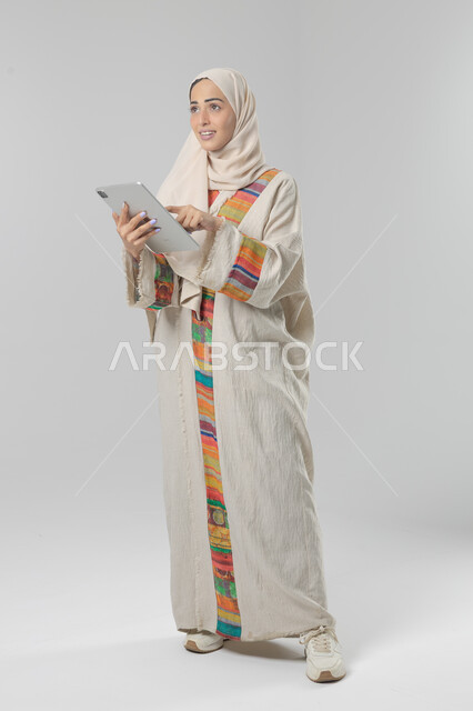  استخدام التقنية الحديثه المتطورة في الحياة العملية عبر الجهاز اللوحي، تعابير وايماءات الاندماج، بورتريه لشابة عربية خليجية سعودية مبتسمة ترتدي الحجاب والعباءة تقوم بالعمل على جهازالتابلت، صورة الجسم بالطول الكامل، خلفية بيضاء 