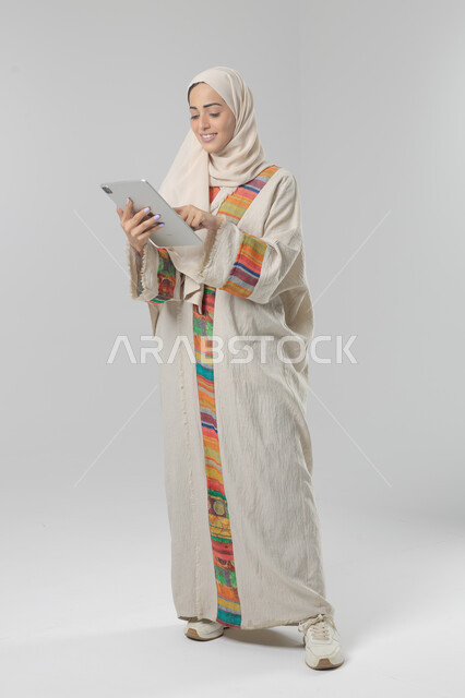 Using modern advanced technology in practical life via tablet, expressions and gestures of integration, portrait of a smiling young Saudi Arabian Gulf woman wearing hijab and abaya working on a tablet, full body image, white background