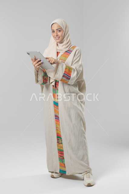 Using modern advanced technology in practical life via tablet, expressions and gestures of integration, portrait of a smiling young Saudi Arabian Gulf woman wearing hijab and abaya working on a tablet, full body image, white background
