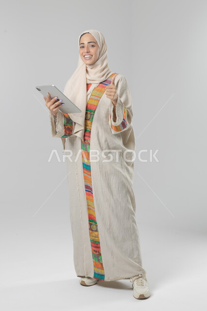  استخدام التقنية الحديثه المتطورة في الحياة العملية عبر الجهاز اللوحي، تعابير وايماءات الاندماج، بورتريه لشابة عربية خليجية سعودية مبتسمة ترتدي الحجاب والعباءة تقوم بالعمل على جهازالتابلت، صورة الجسم بالطول الكامل، خلفية بيضاء 