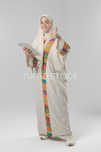 Using modern advanced technology in practical life via tablet, expressions and gestures of integration, portrait of a smiling young Saudi Arabian Gulf woman wearing hijab and abaya working on a tablet, full body image, white background