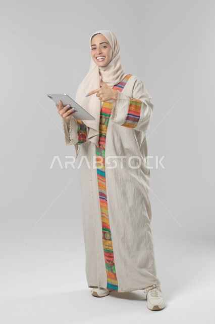  استخدام التقنية الحديثه المتطورة في الحياة العملية عبر الجهاز اللوحي، تعابير وايماءات الاندماج، بورتريه لشابة عربية خليجية سعودية مبتسمة ترتدي الحجاب والعباءة تقوم بالعمل على جهازالتابلت، صورة الجسم بالطول الكامل، خلفية بيضاء 