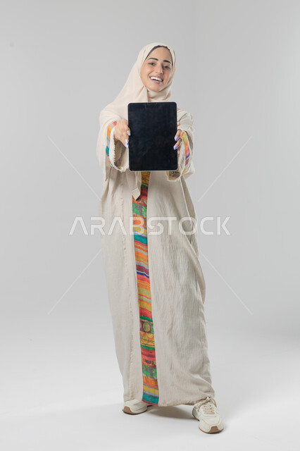 Tablet screen display, use of modern advanced technology in practical life, expressions and gestures of integration, portrait of a smiling young Saudi Arabian Gulf woman wearing hijab and abaya working on a tablet, full body image, white background